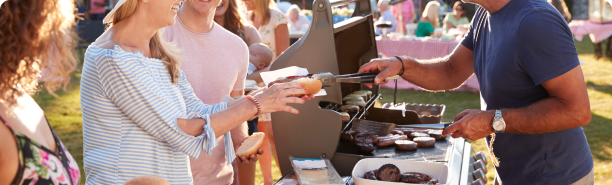 A person serving barbecued hot dogs and hamburgers to a line of guests at an outdoor party.