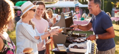 A person serving barbecued hot dogs and hamburgers to a line of guests at an outdoor party.