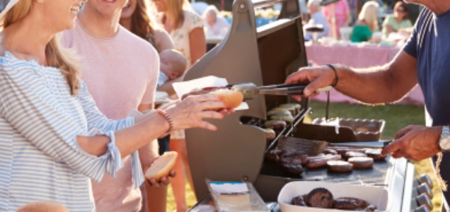 A person serving barbecued hot dogs and hamburgers to a line of guests at an outdoor party.